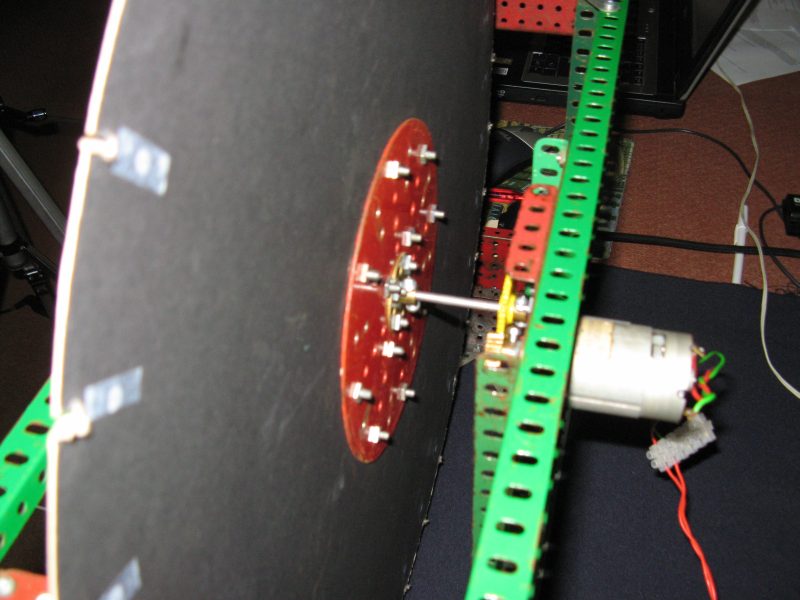 Figure 5 — A Meccano-framed NBTV disc televisor mid-build, showing the small DC motor coupled to a red LED-cluster mounting plate on the disc's central hub, with the black disc itself visible edge-on to the l…