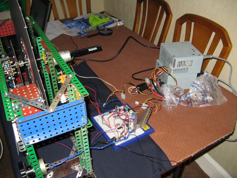 Figure 6 — A hobbyist NBTV build's electronics laid out on a bench: a small breadboard-mounted sync-separator/motor-control circuit, wiring looms to the Meccano-framed disc-and-motor assembly, and a switched-…