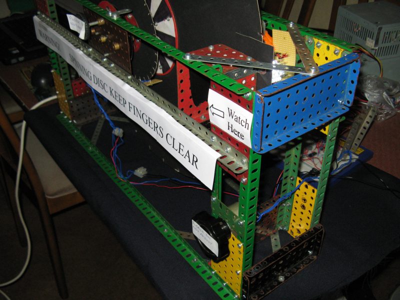 Figure 1 — A member-built NBTV disc televisor, framed in Meccano, carrying a hand-lettered warning label reading "SPINNING DISC KEEP FINGERS CLEAR" with a "Watch Here" arrow pointing to the safe viewing posit…